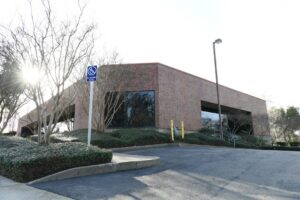 Brick office building with large windows, planted landscaping, and a blue wheelchair-accessible sign in the parking area; sun glare through bare trees.